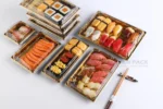 Assorted sushi containers with clear lids holding sushi rolls, sashimi, and sliced fish
