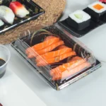 Salmon sushi arranged in a black WL-03 tray with a transparent lid in a dining setting
