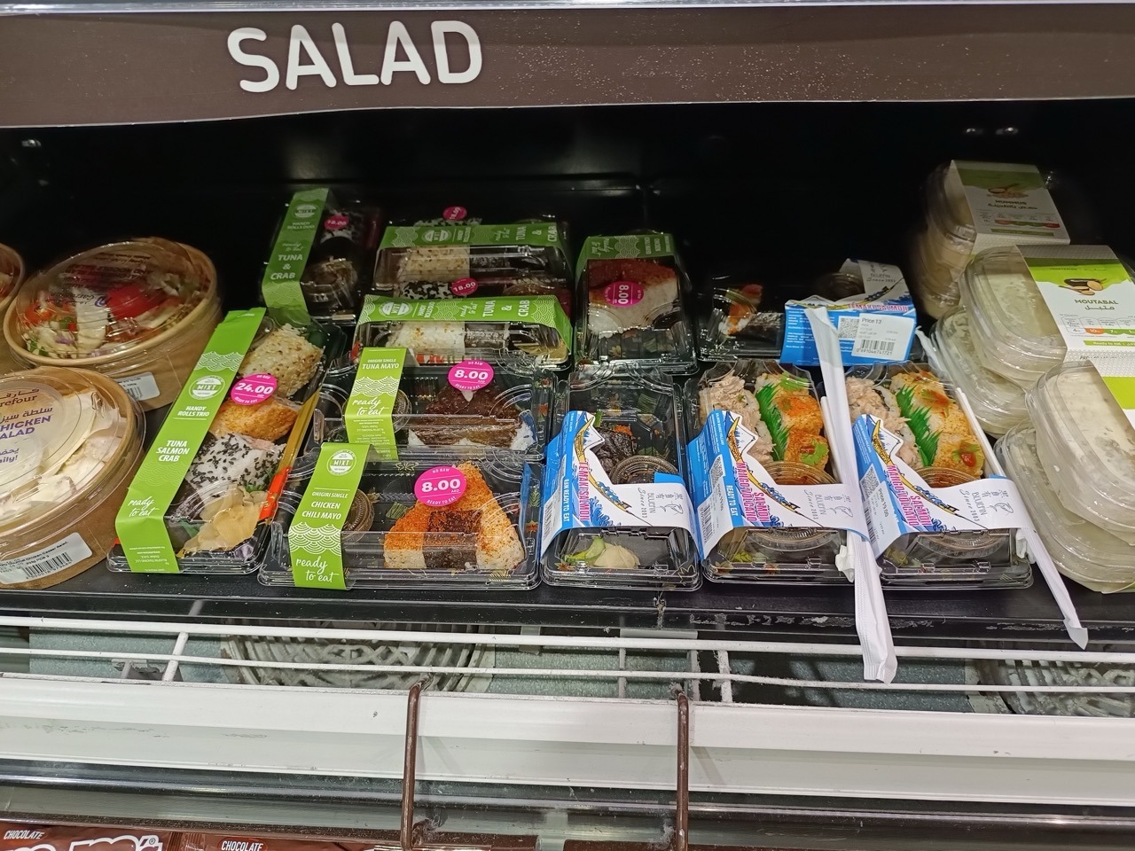Clear plastic food packaging trays containing ready-to-eat salads, sushi and prepared meals displayed in a refrigerated retail case.